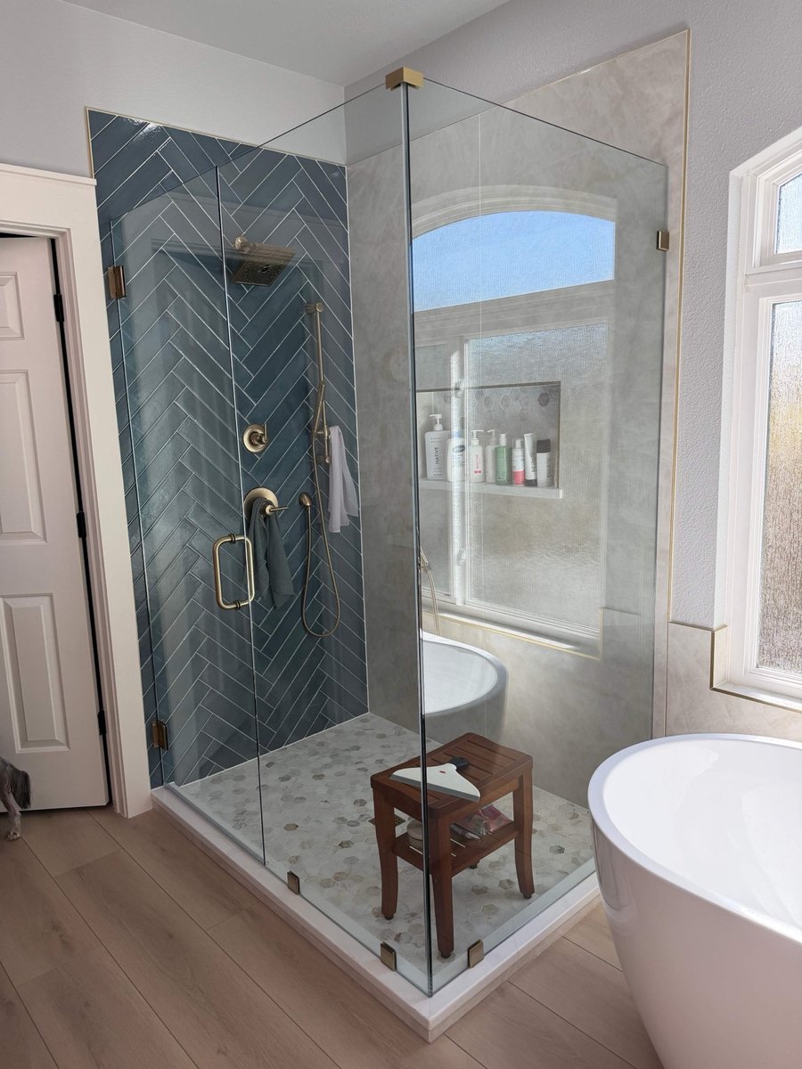 Corner glass shower with blue herringbone wall and teak stool beside freestanding tub