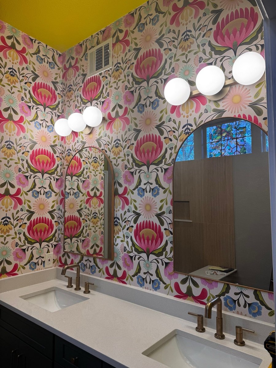 Double vanity with bold floral wallpaper, arched mirrors, and brass faucets