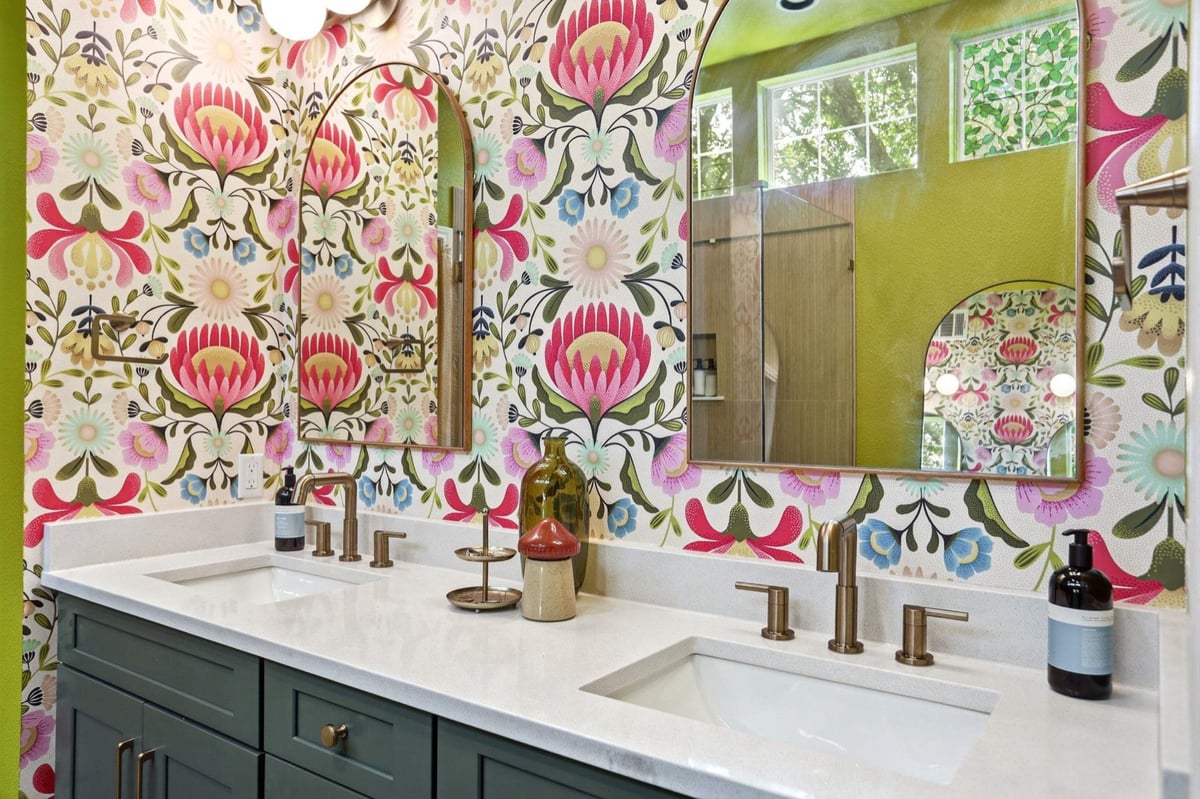 Twin arched mirrors and brass faucets over a teal vanity with bold wallpaper