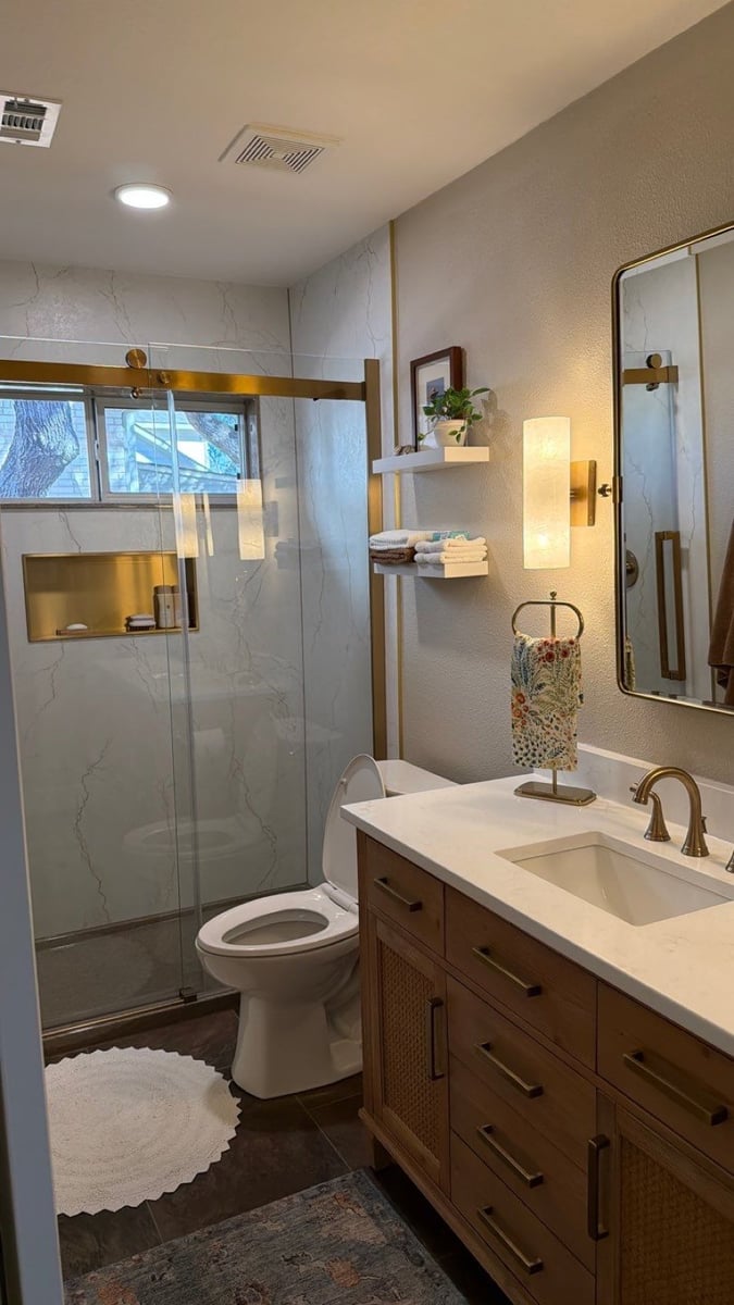 Remodeled bathroom with glass walk-in shower, marble walls, wood vanity and gold fixtures