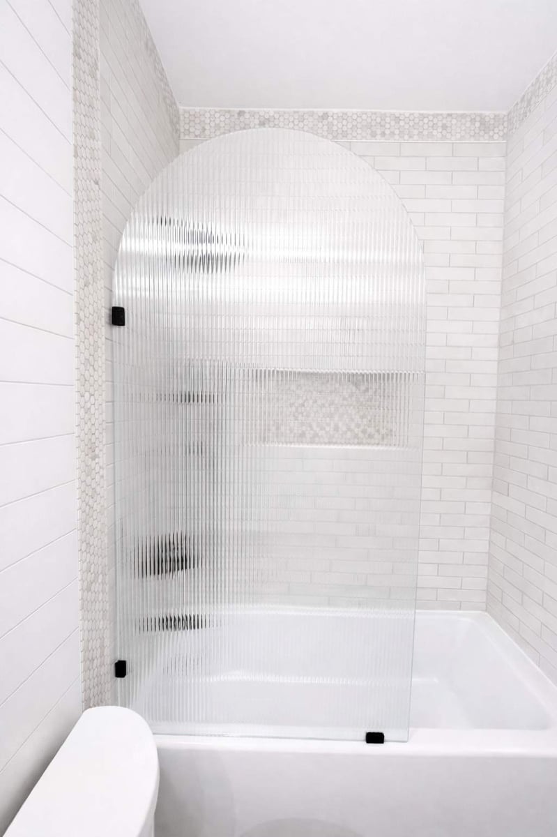 Arched fluted glass screen on tub framed by white subway and penny tile