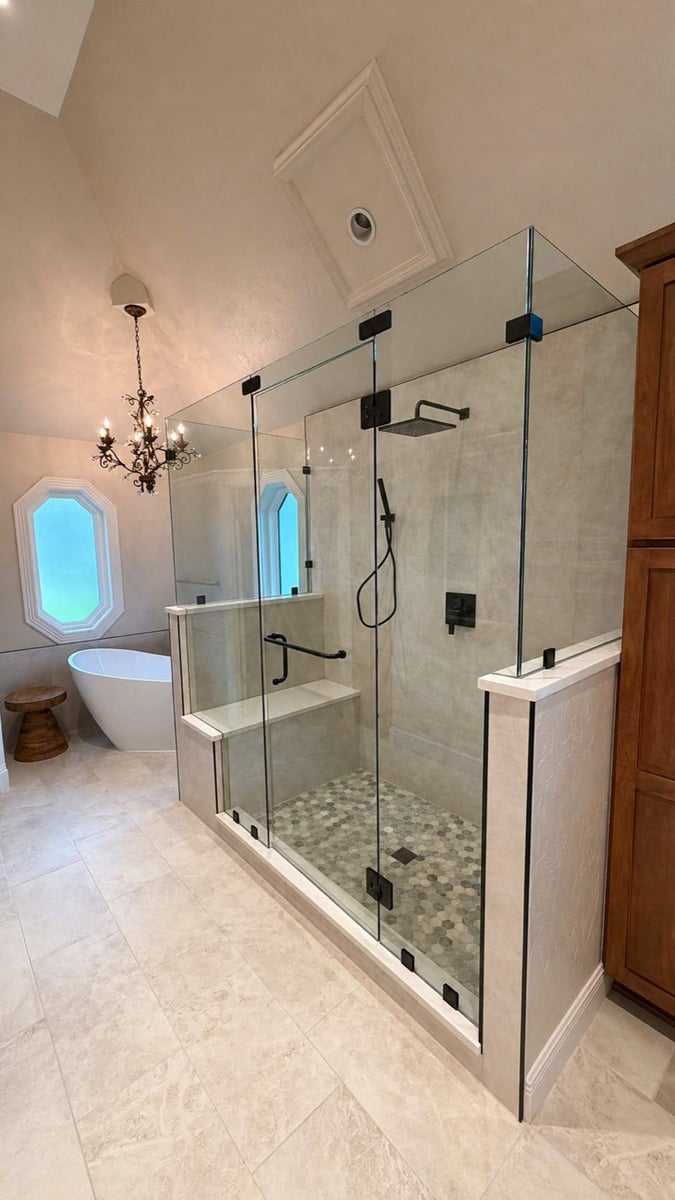 Large frameless glass shower with black fixtures beside freestanding tub and chandelier