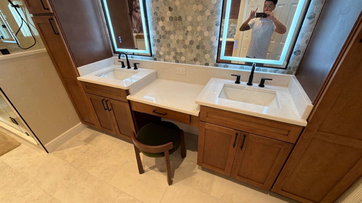 Double vanity with backlit mirrors, wood cabinets, seated makeup station, and green stool