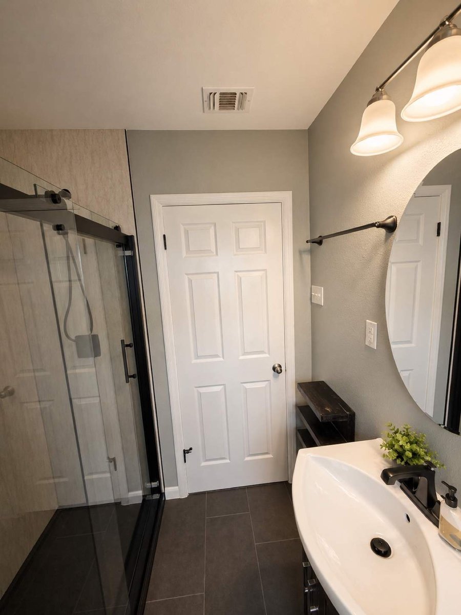 Sliding glass shower with marble-look walls and oval sink under round mirror