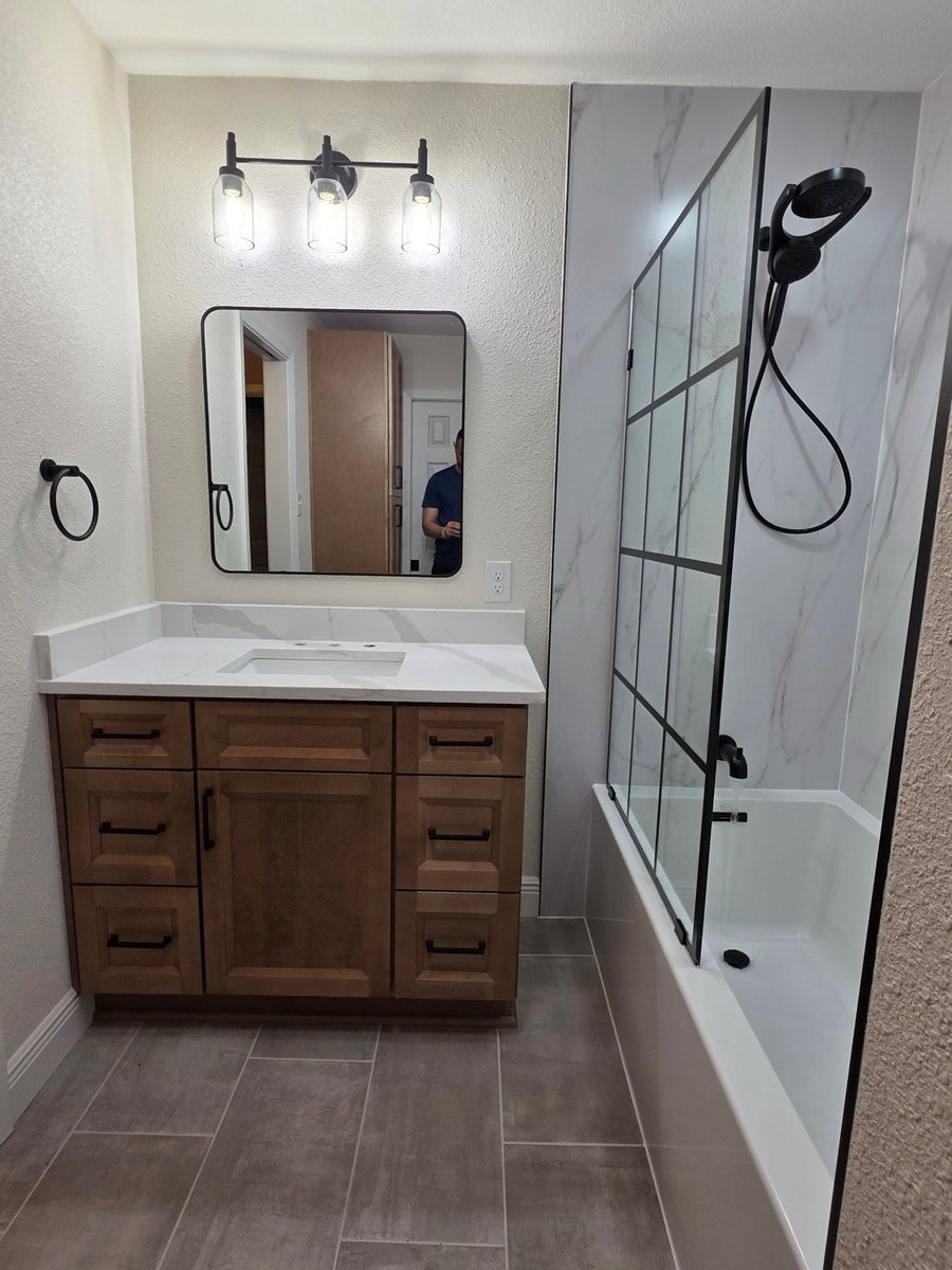 Wood vanity with white quartz top under black-framed mirror beside tub-shower combo