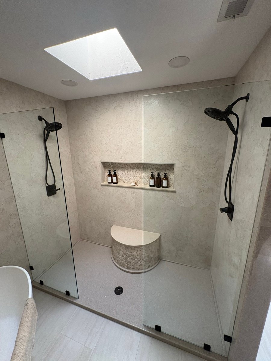 Glass walk-in shower with beige stone walls, curved bench and black fixtures under skylight