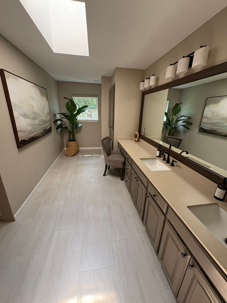 Long double vanity with taupe cabinets, dark framed mirror and tile floor below skylight