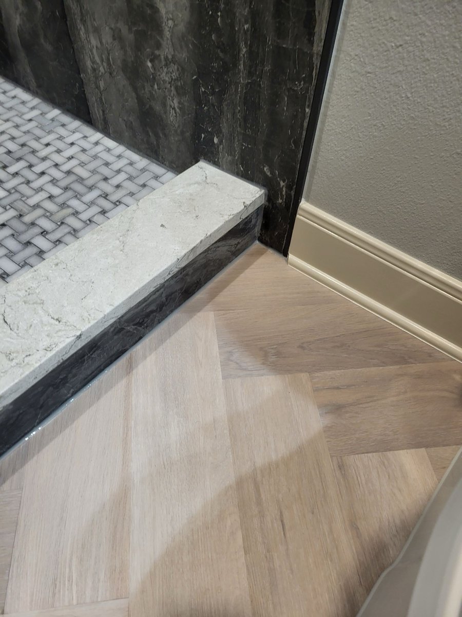 Detail of dark marble shower curb meeting herringbone wood-look floor tile