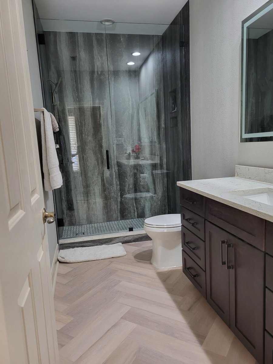 Walk-in glass shower in gray stone beside dark vanity and herringbone floor