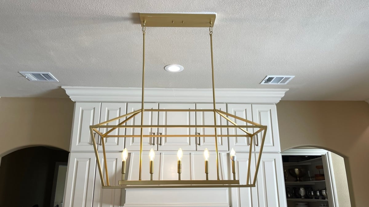 Brass linear cage chandelier hung below crown molding on cream cabinets