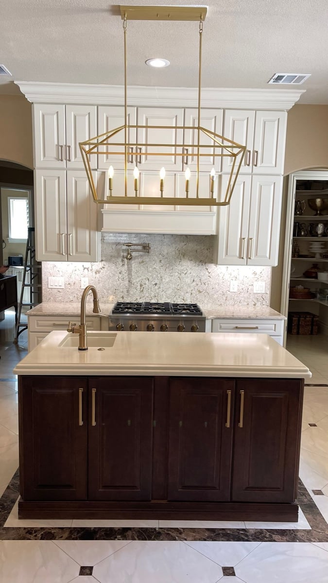 Dark island with gold faucet under brass pendant facing gas range and white cabinets