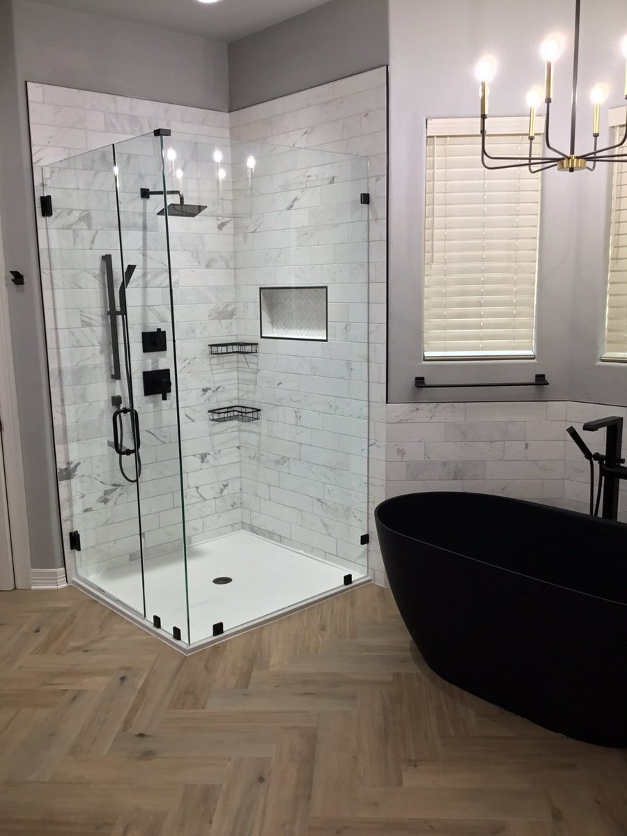 Glass shower with marble tile and black tub under brass chandelier
