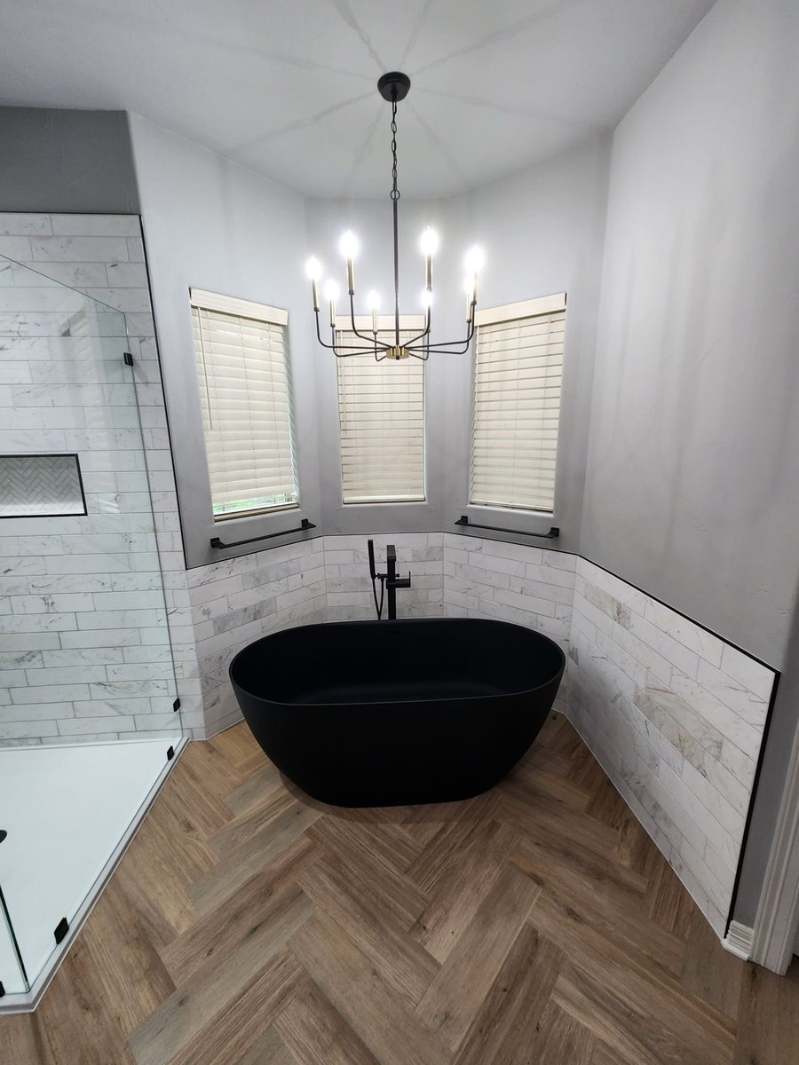 Black freestanding tub centered under chandelier between three blind windows