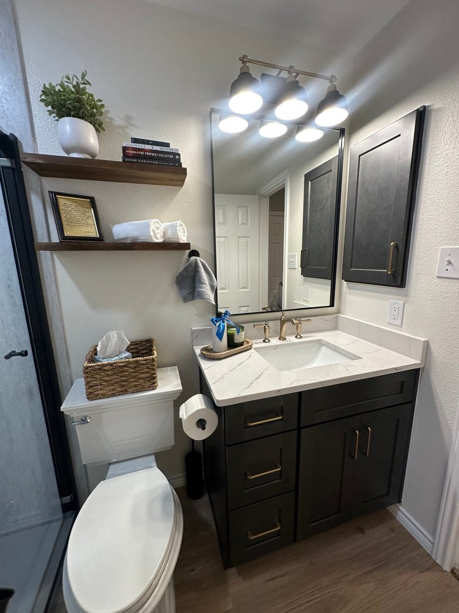Dark vanity with white quartz top, framed mirror, floating wood shelves, and gold fixtures