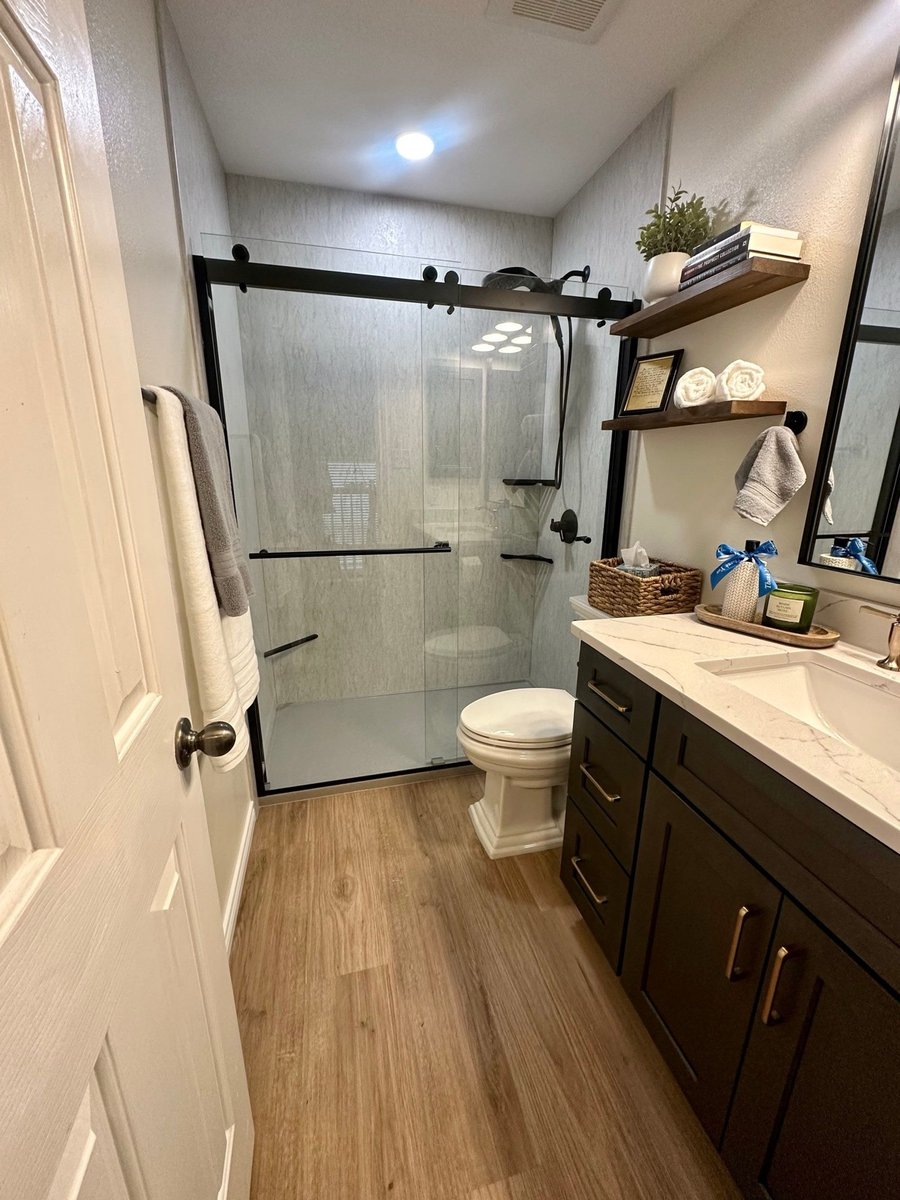 Wide view of remodeled bathroom with glass shower, dark vanity, and wood-look floor