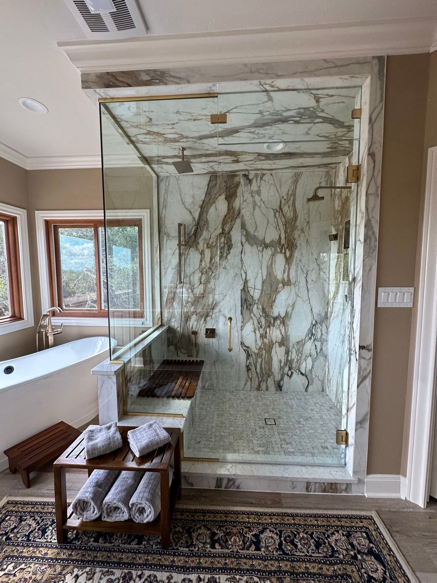 Marble shower with frameless glass beside a freestanding tub and teak bench