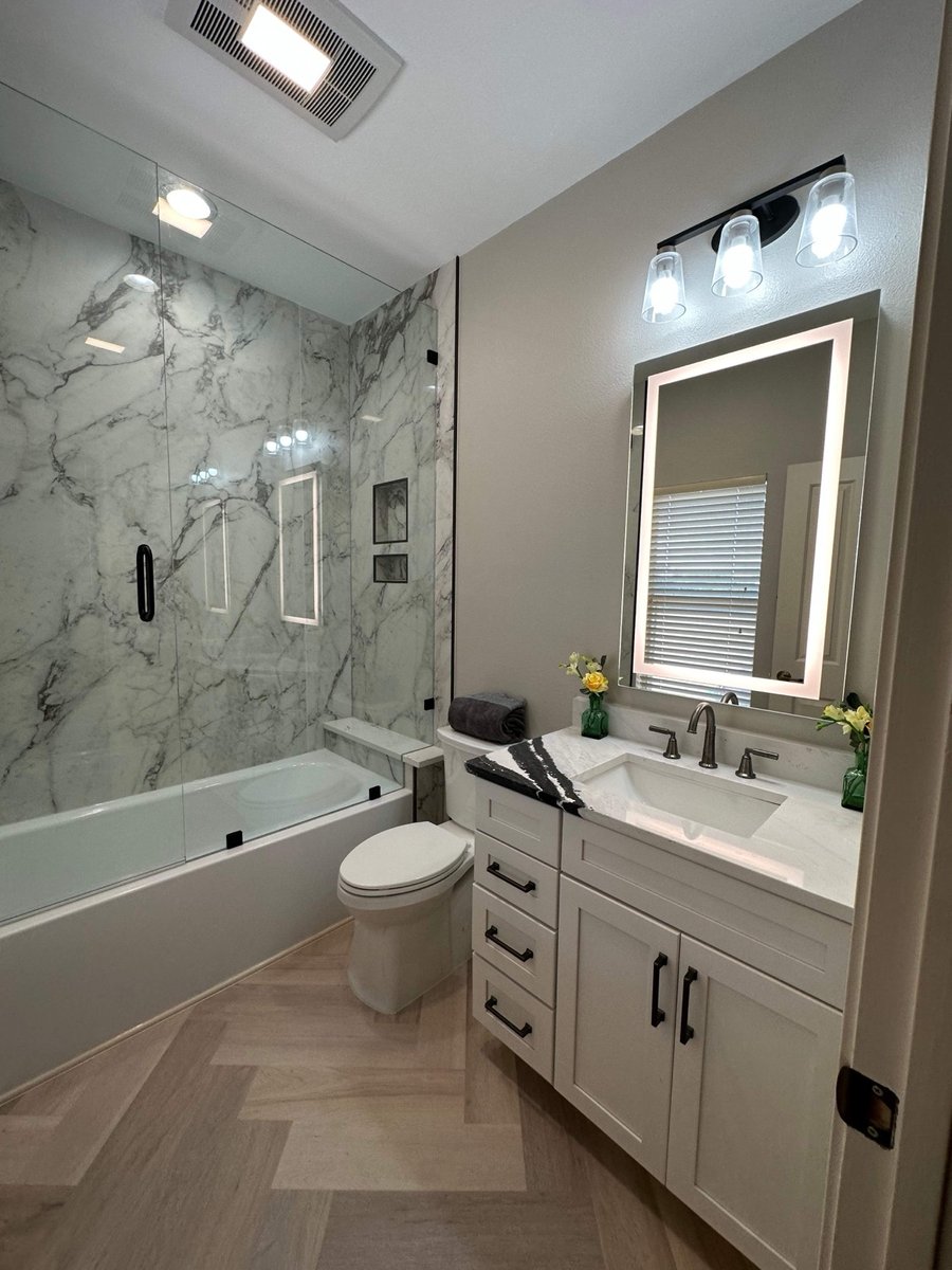 Marble tub-shower combo with glass enclosure beside lit mirror and white vanity