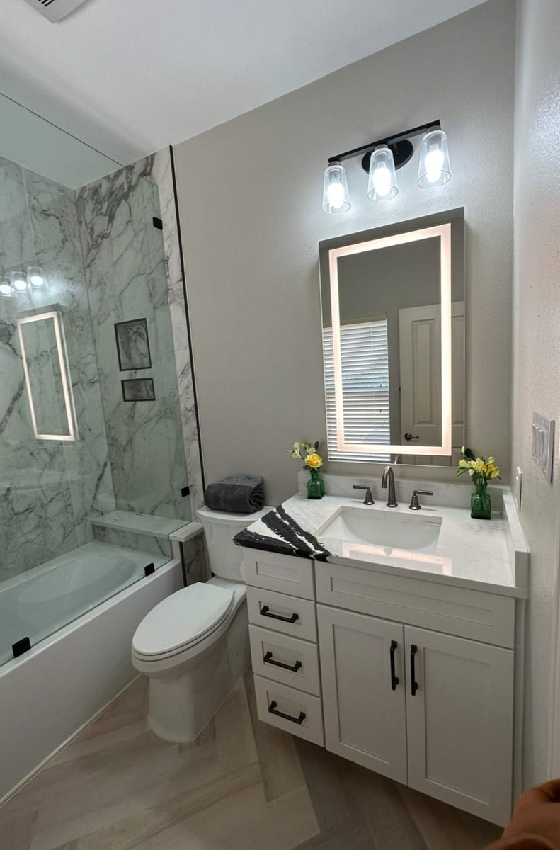 Compact bath with marble tub surround, lighted mirror, and white vanity with yellow flowers