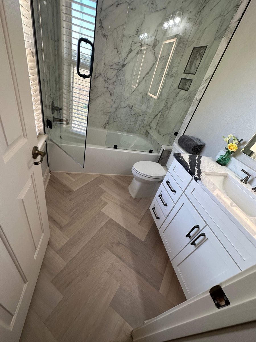 Marble tub shower, toilet, and white vanity over herringbone wood-look tile
