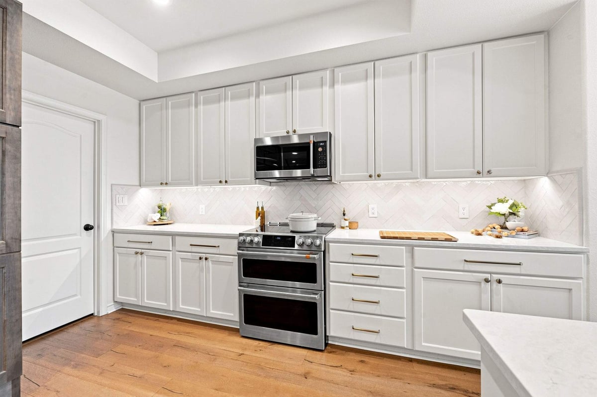 White Shaker kitchen cabinets with brass hardware, stainless steel range, and herringbone tile backsplash.