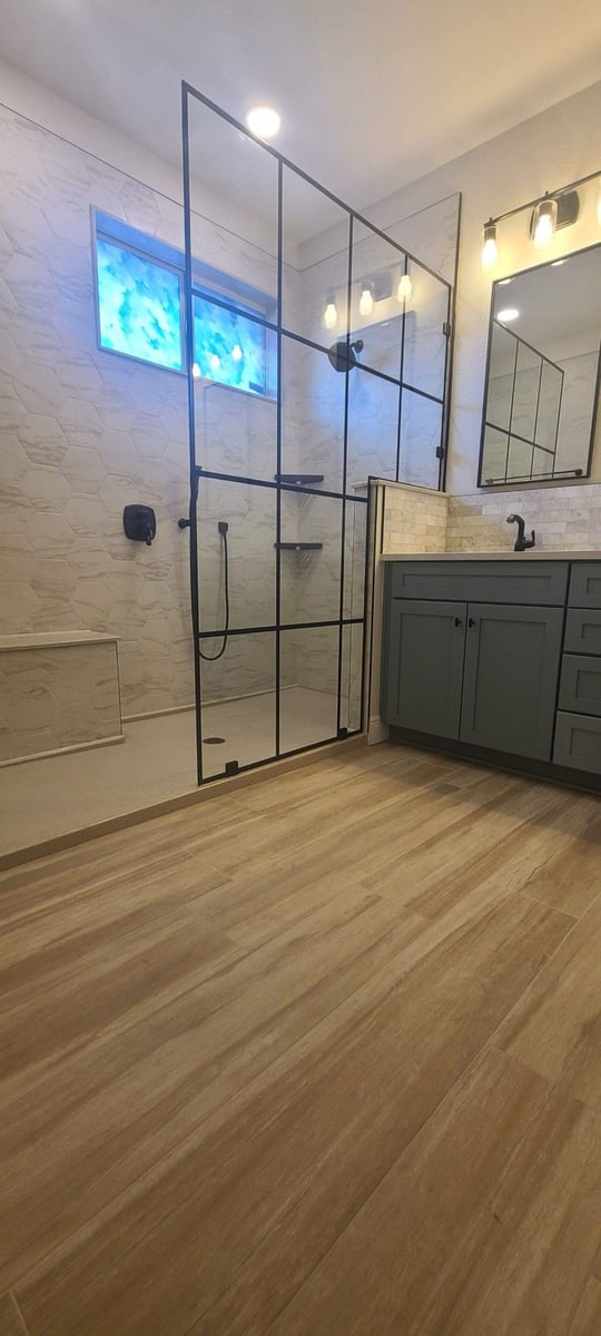 Glass shower with black grid framing beside wood-look tile floor and green vanity