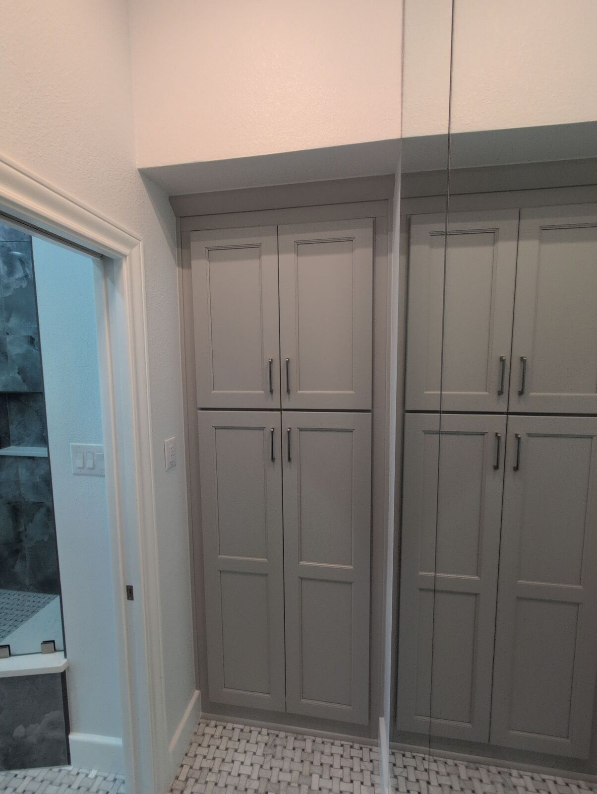 Bathroom storage cabinets in warm gray with paneled doors, mirrored wall and mosaic tile floor.