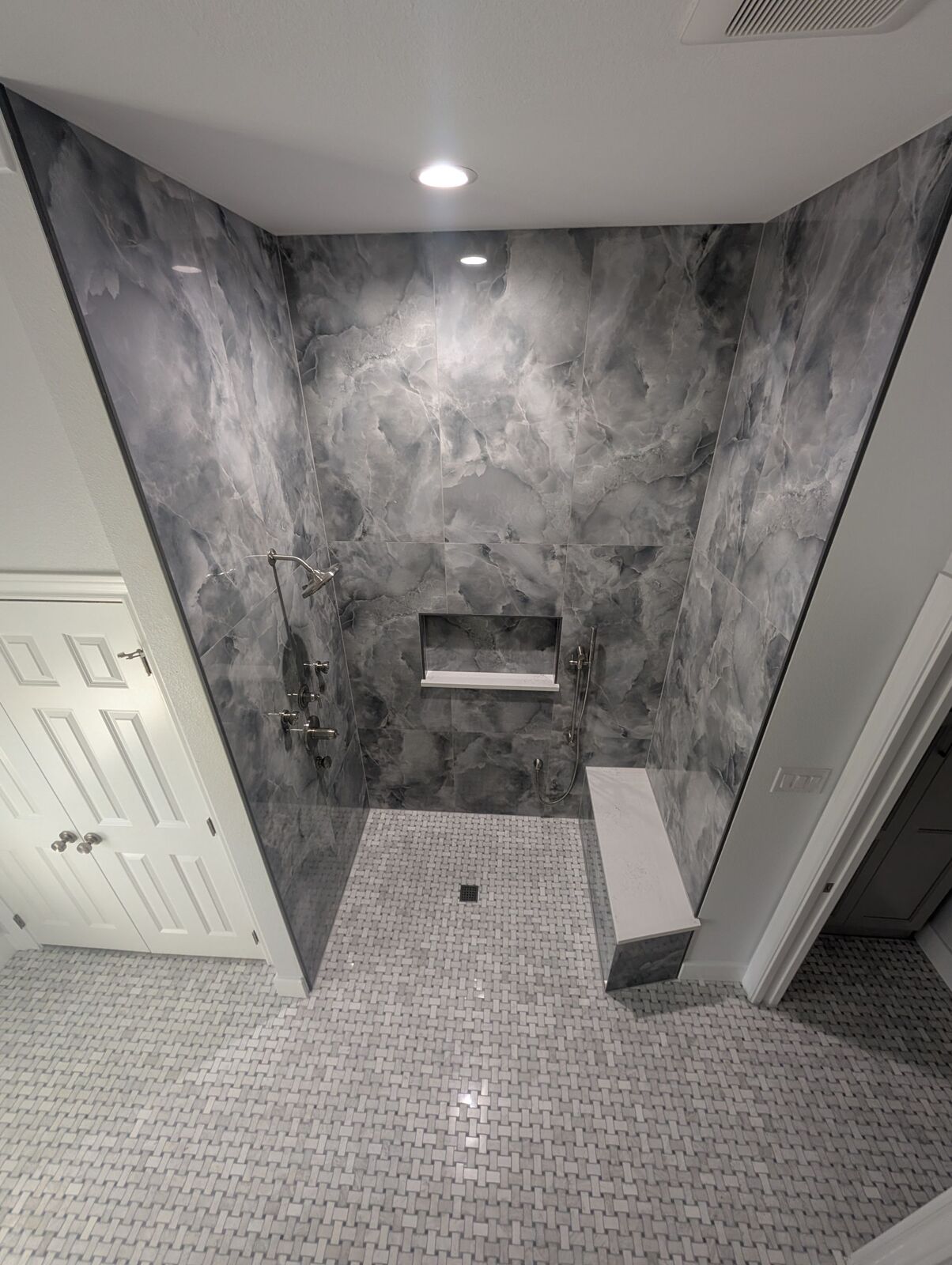 Walk-in shower with dark gray marble-look tile, glass panels, bench, niche and mosaic bathroom floor.